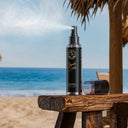 SHARK SURF Sea Salt Spray bottle with cap off showing spray effect on a wooden table on a beach with the ocean in the background